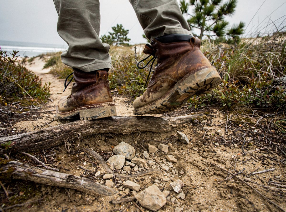 A pair of worn hiking boots step over exposed roots and loose stones on a trail